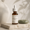 An amber glass dropper bottle labeled 'Onise. Peptide Hair Growth Serum' stands on a light stone slab beside a green soap bar, with a large textured vase and soft shadowy leaves in the background.