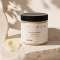 A jar of Onise Raw Shea Butter with vitamins A&E sits on a textured stone slab with a smear of the butter beside it. The scene is styled with warm, natural shadows.